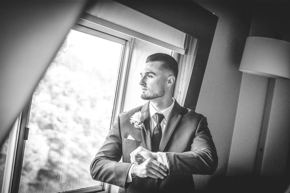 Black and white portrait of a handsome groom in a suit adjusting his cuffs by a bright window.