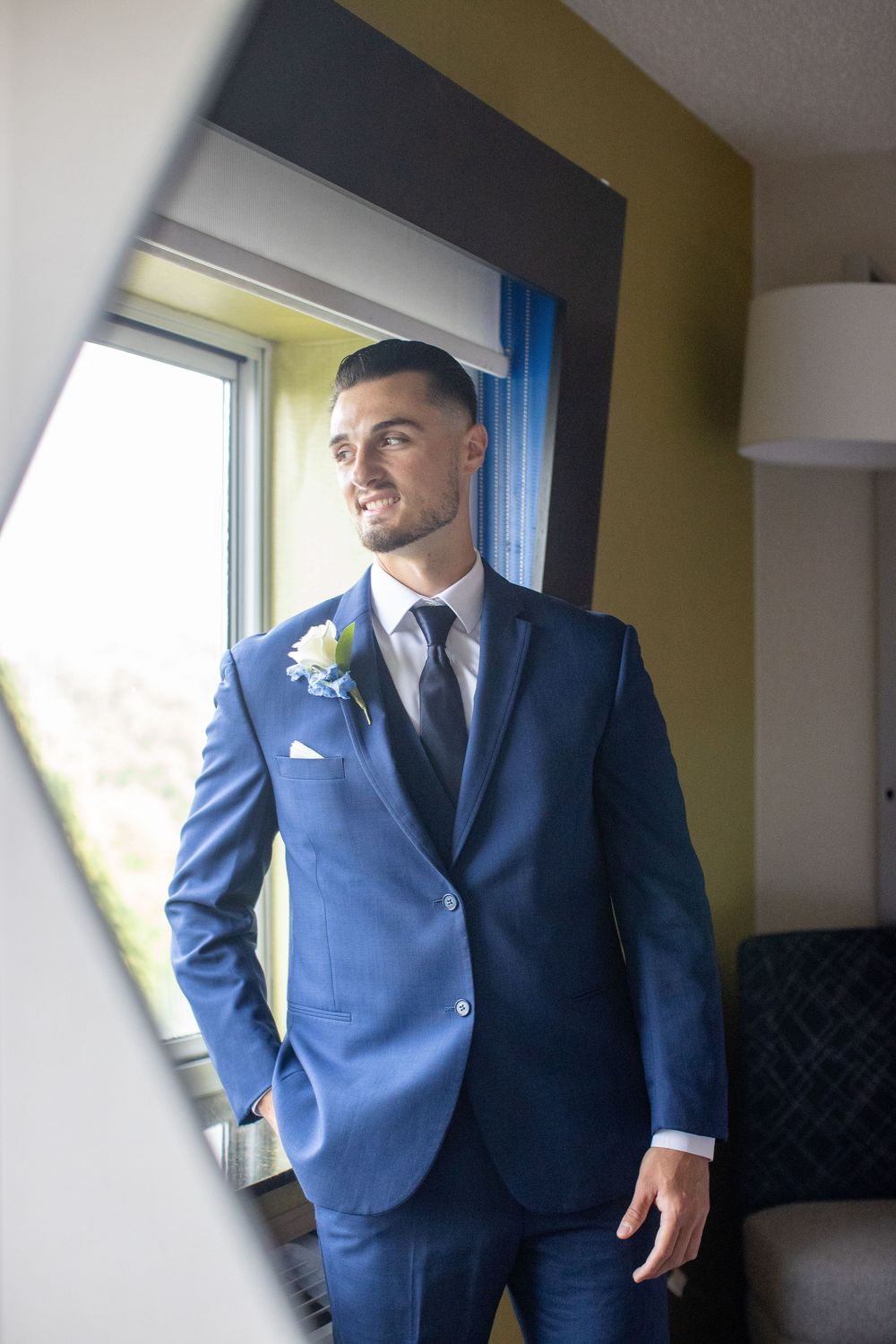 Dapper groom in navy blue suit with boutonniere smiles while standing by a window in a hotel room.