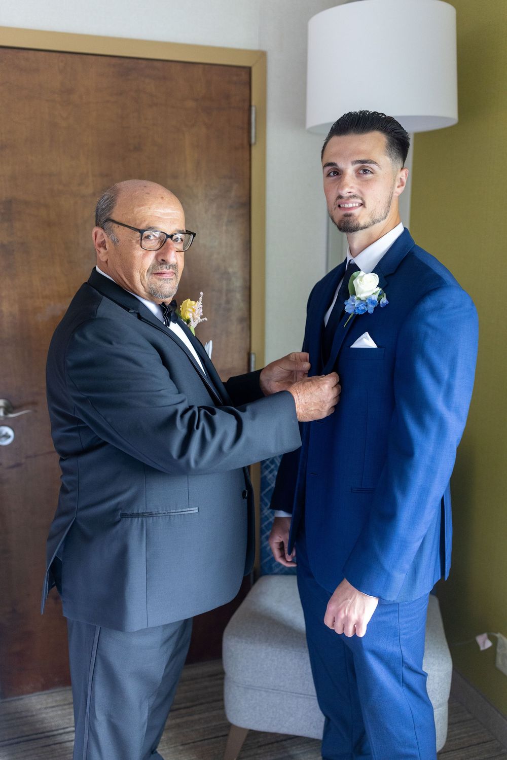 Older man helping groom adjust boutonniere on blue suit before wedding ceremony.