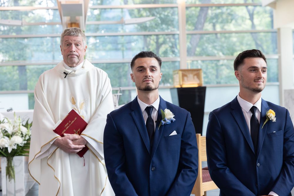 Groom and groomsman standing at altar in blue suits with priest during wedding ceremony.