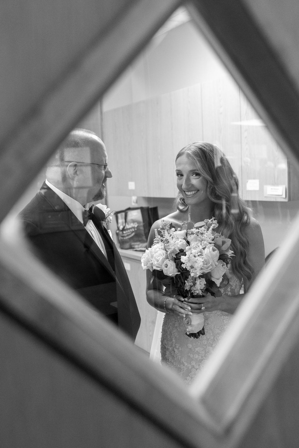 Bride smiling with bouquet alongside father reflected through diamond-shaped window frame.