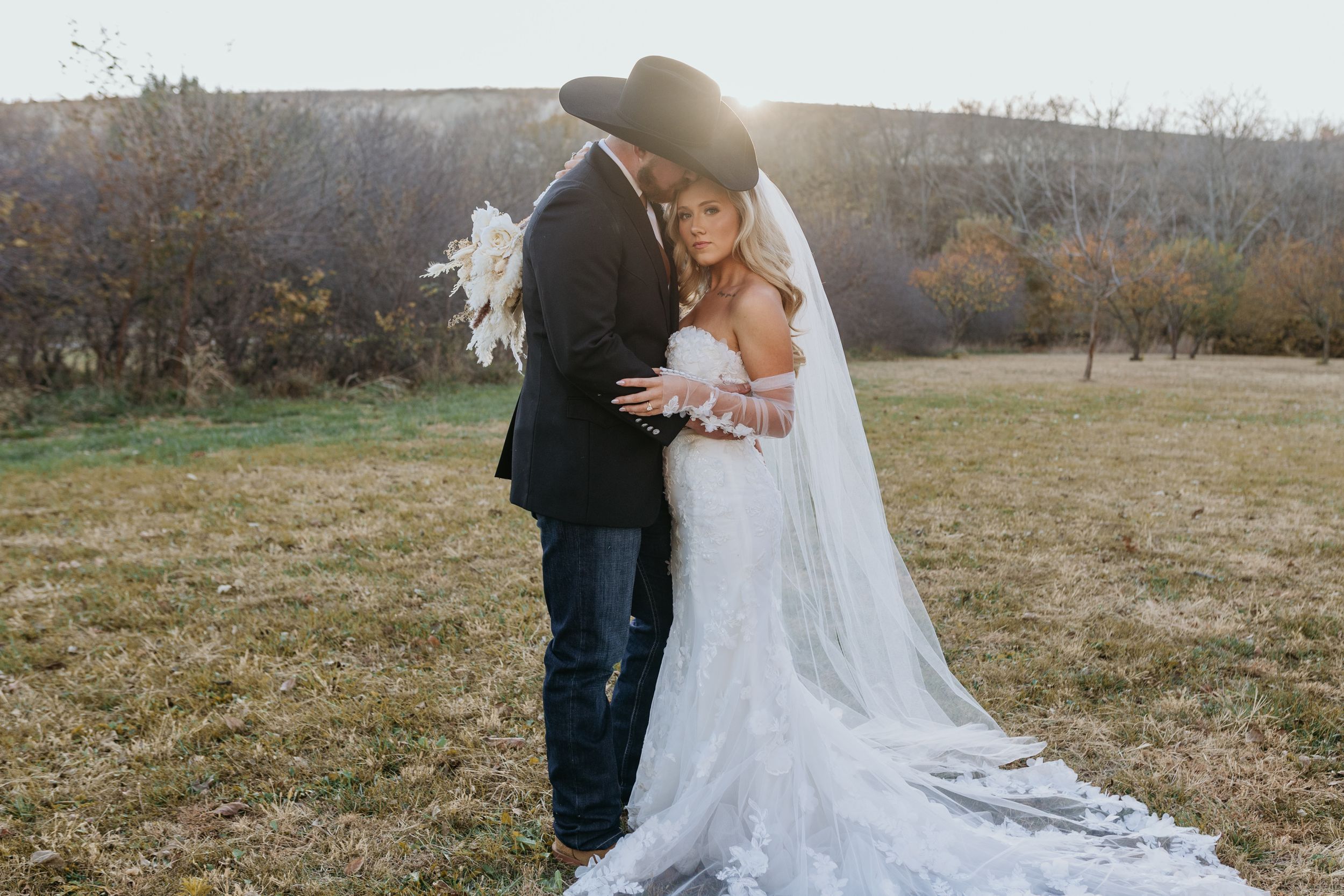 Couple in western-inspired attire embraces during fall wedding ceremony with long flowing white dress and veil in field.