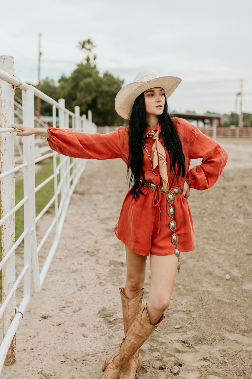 A women in a bright red romper and cowboy boots poses against a white fence at a ranch on an overcast day.