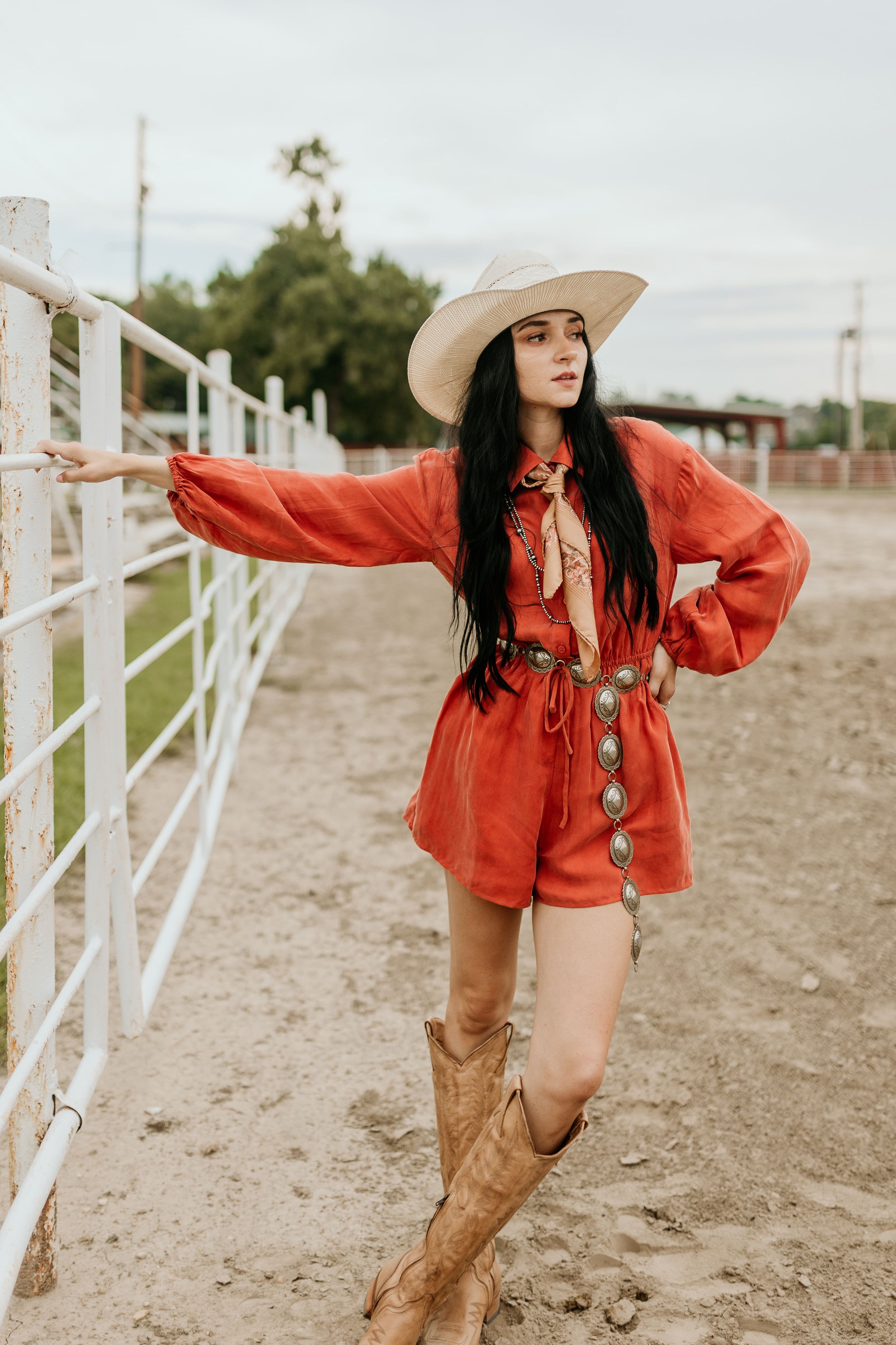 A women in a bright red romper and cowboy boots poses against a white fence at a ranch on an overcast day.