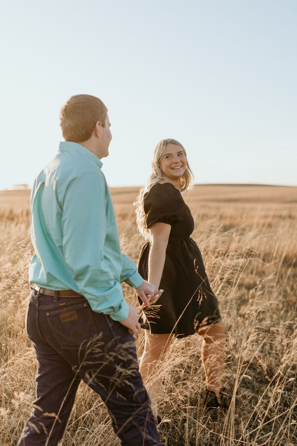 Couple walking through golden field at sunset with woman in black dress looking back and smiling.