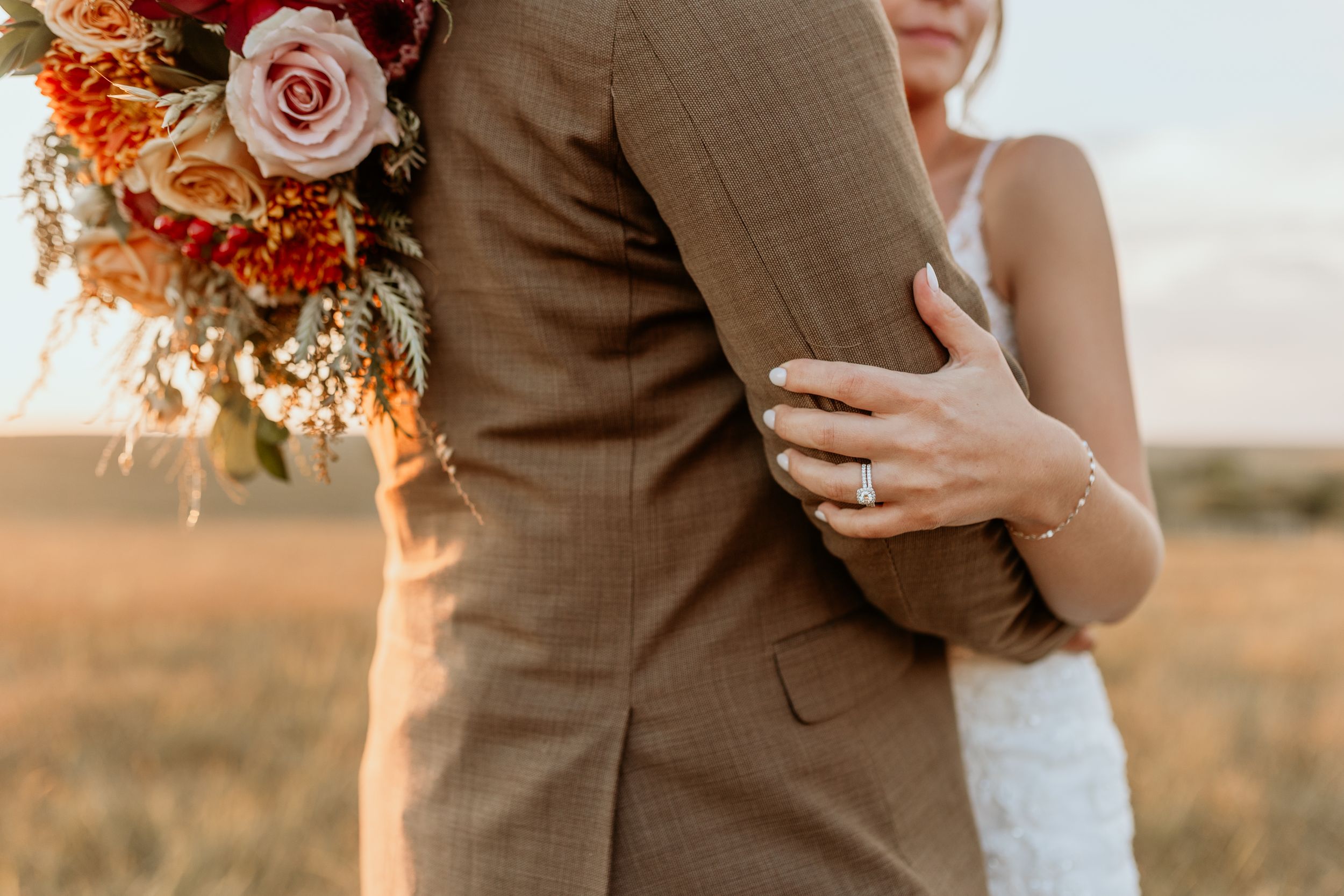 Closeup of a couple embracing in formal attire at sunset with a colorful rose and orange floral bouquet.