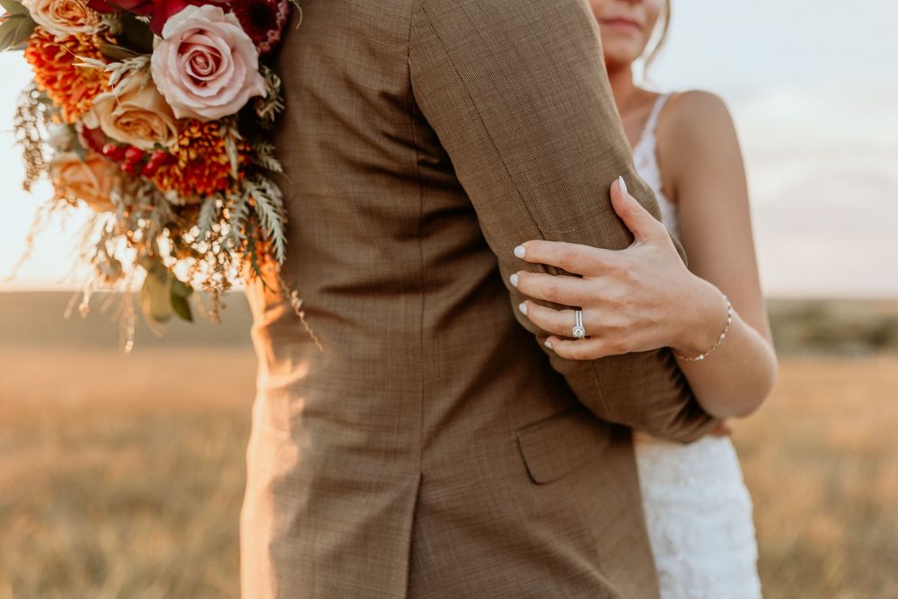 Closeup of a couple embracing in formal attire at sunset with a colorful rose and orange floral bouquet.