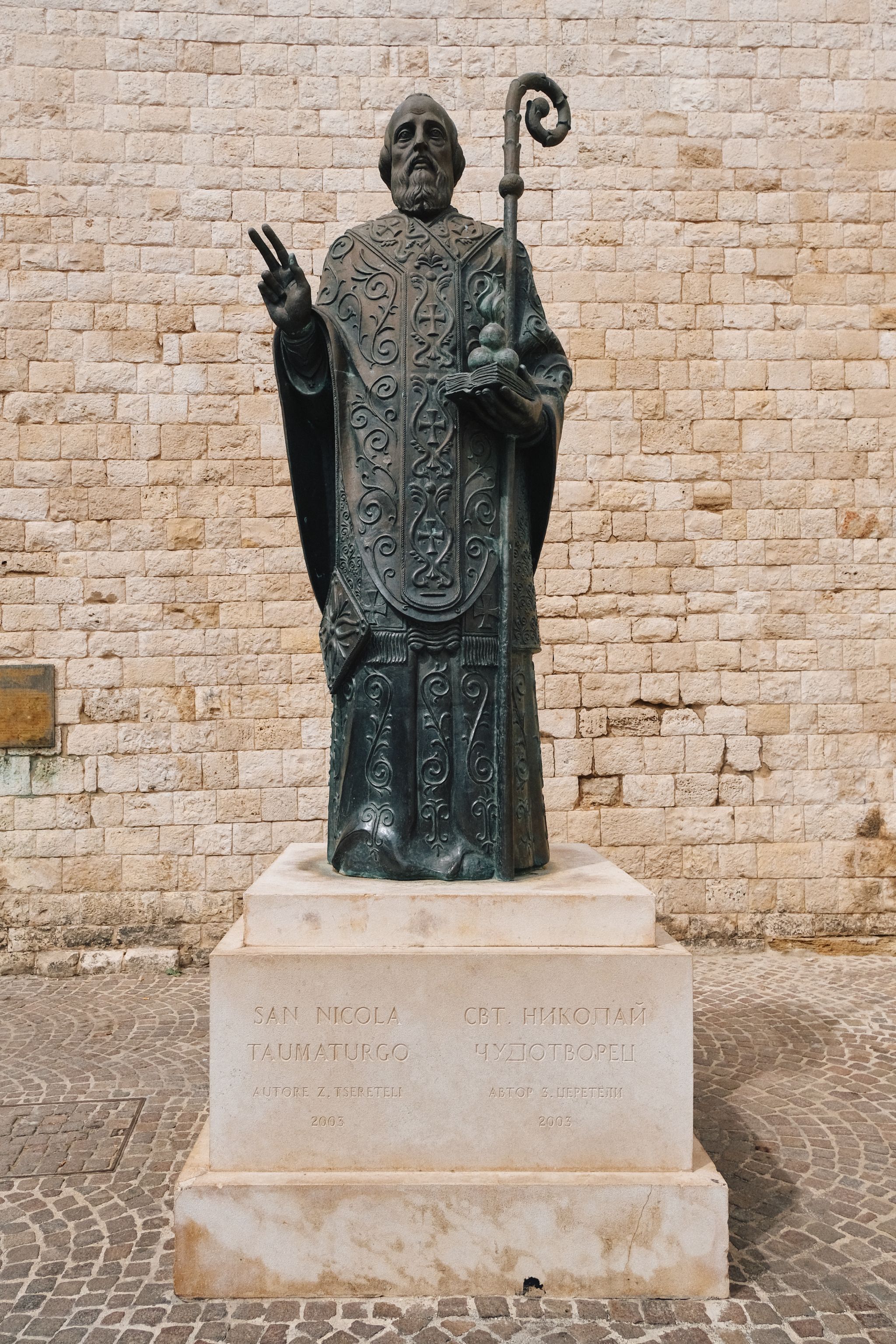 Bronze statue of a religious figure holding a staff stands on stone pedestal in city square.
