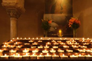 Multiple lit votive candles create a warm glow in a church interior with stone columns and floral arrangements.