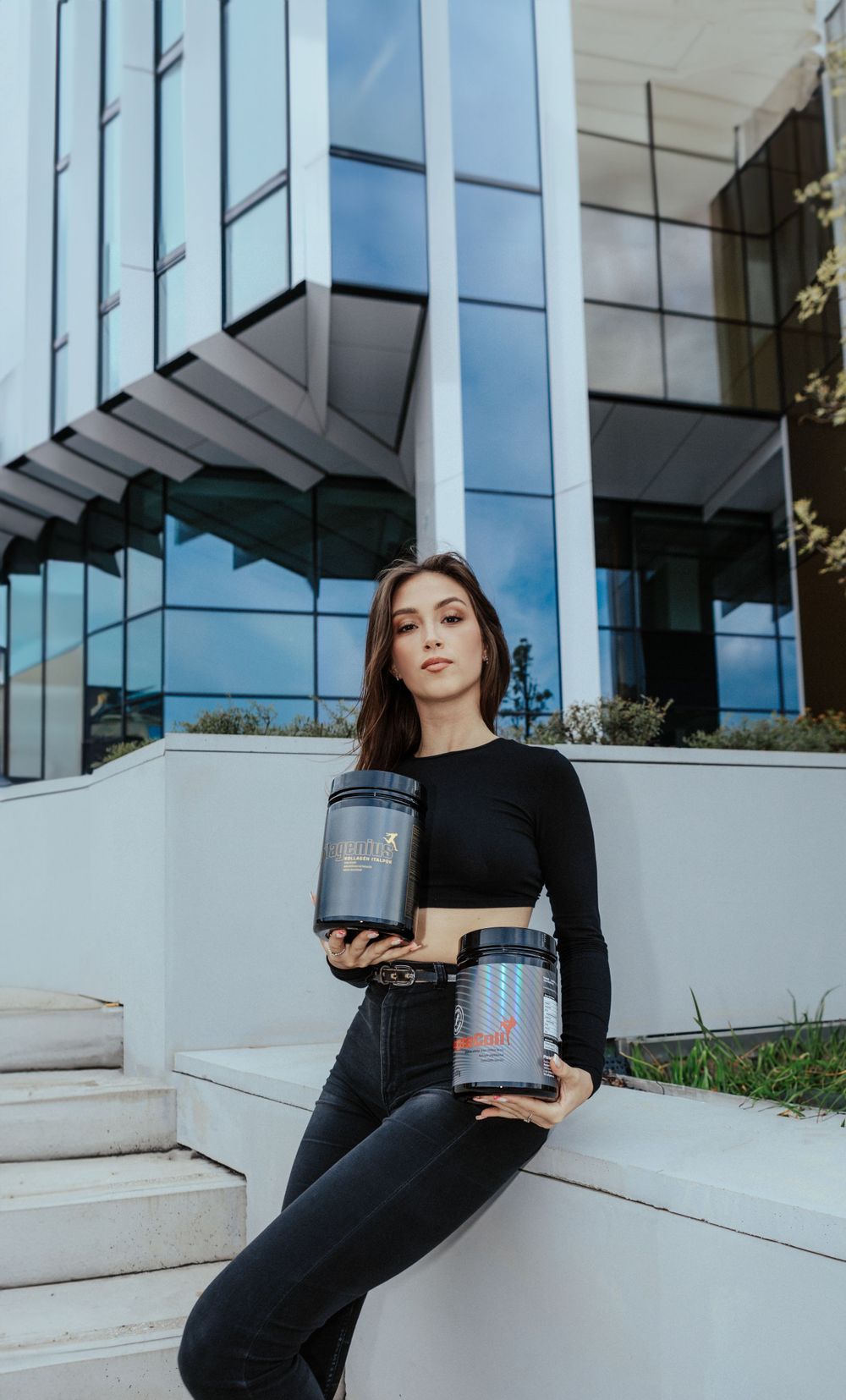 Athletic model in black outfit holding supplement containers against modern glass building.
