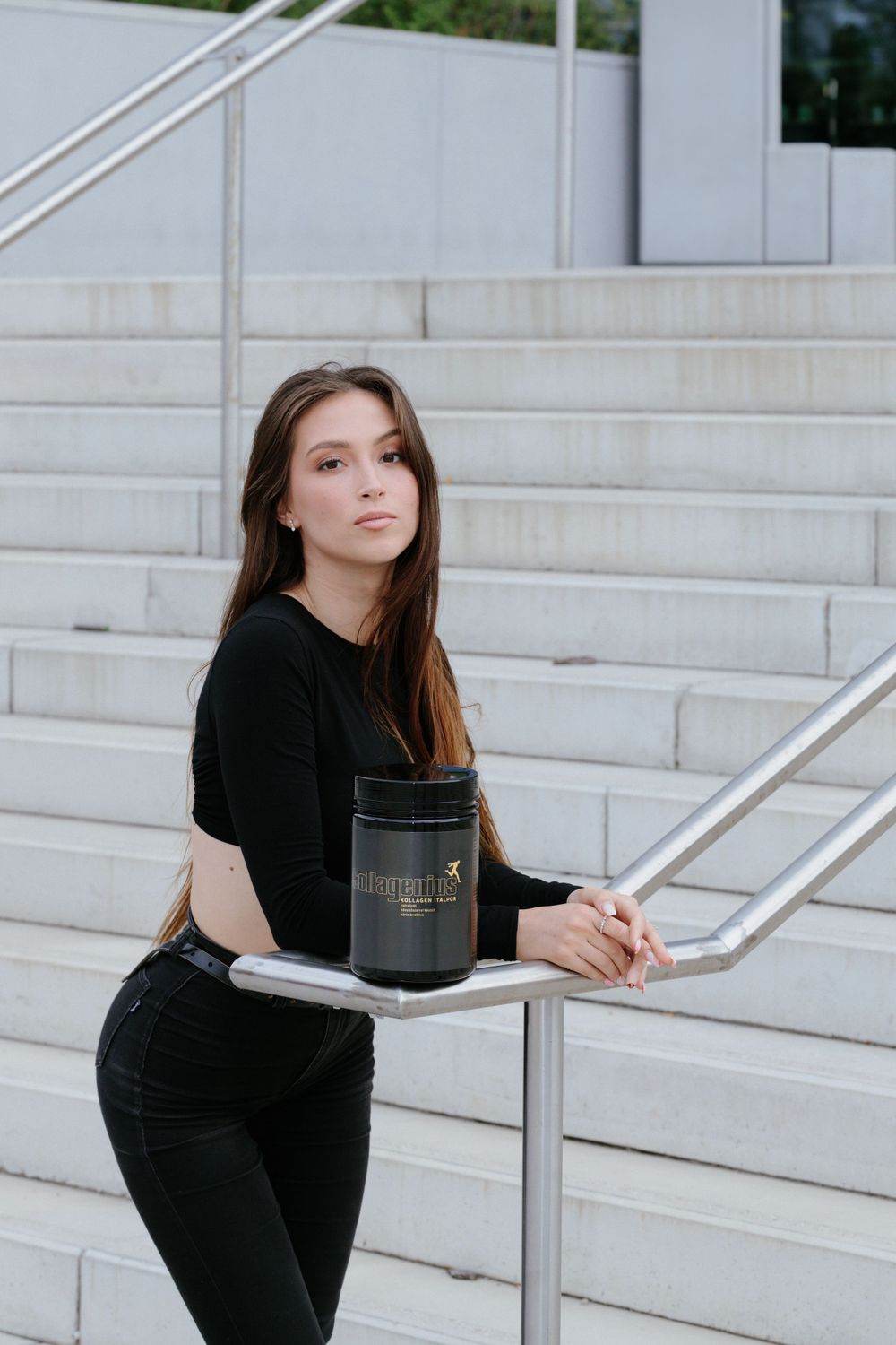 Fitness model in black athletic wear posing with supplement container on white stairs.