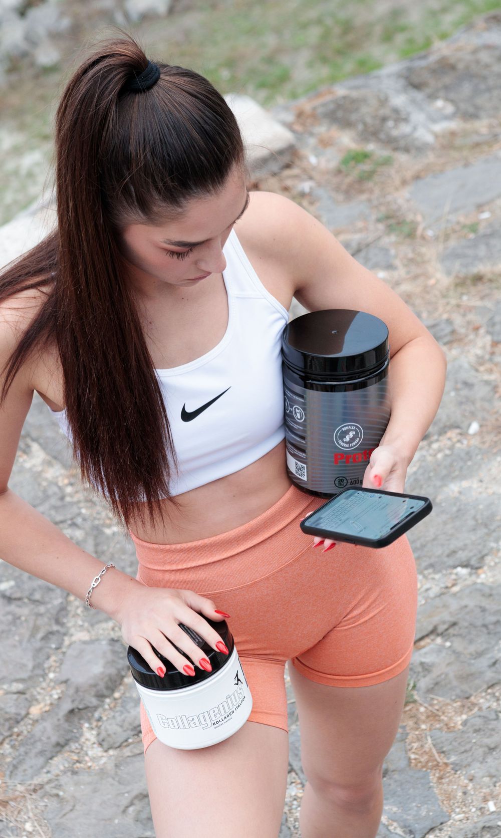Fitness enthusiast in sportswear checking phone while holding supplement containers.