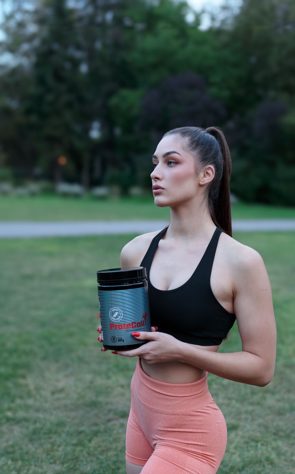 Athletic woman in coral workout shorts holding supplement container in park setting.
