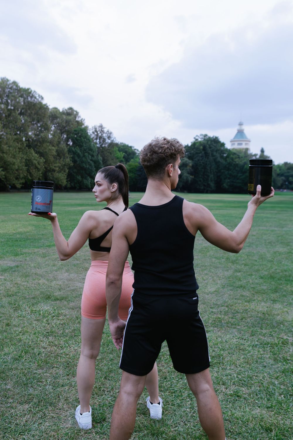 Fitness models displaying supplement containers in park with dome building in background.