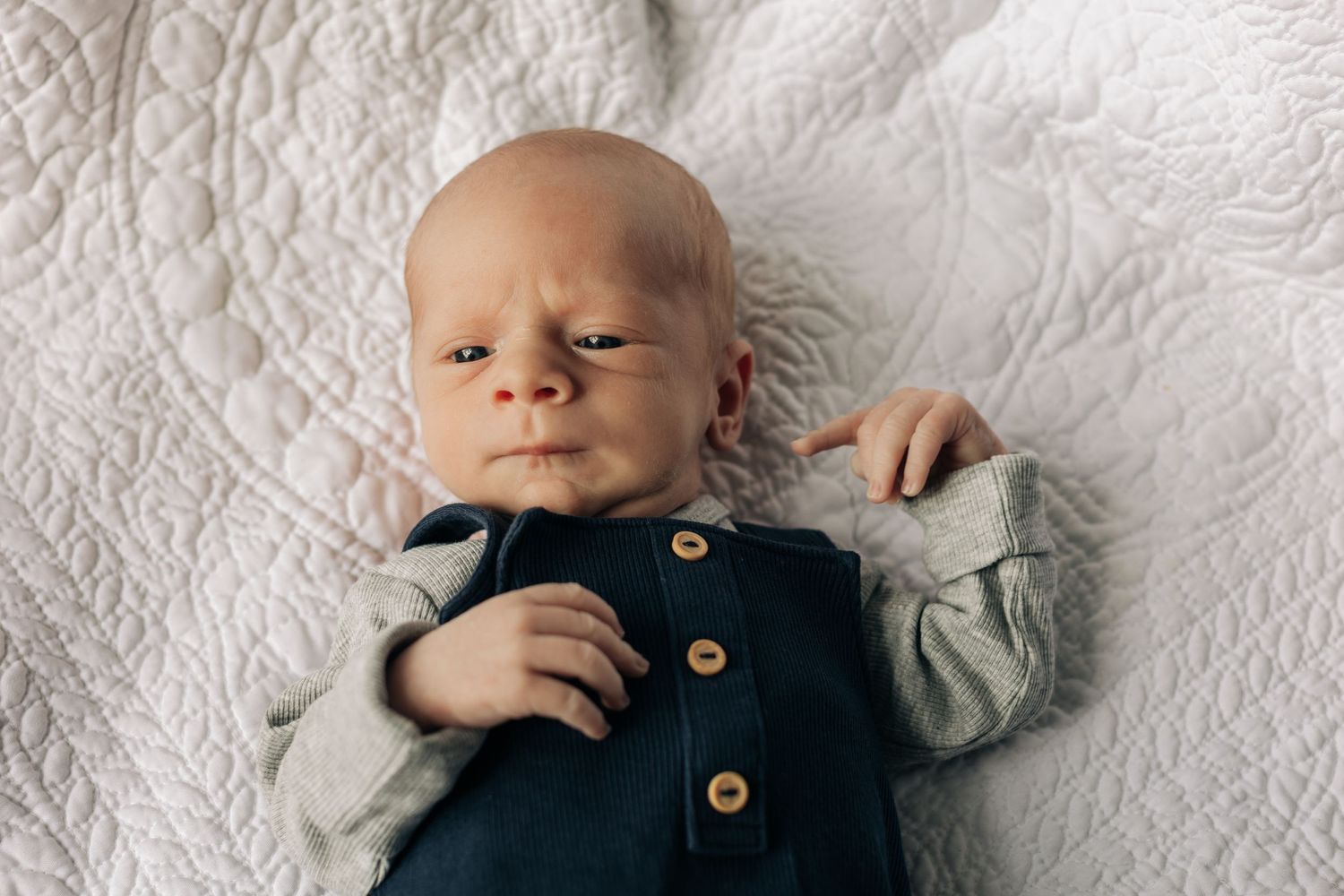 A newborn baby wearing navy overalls with wooden buttons lies on a textured white quilt.