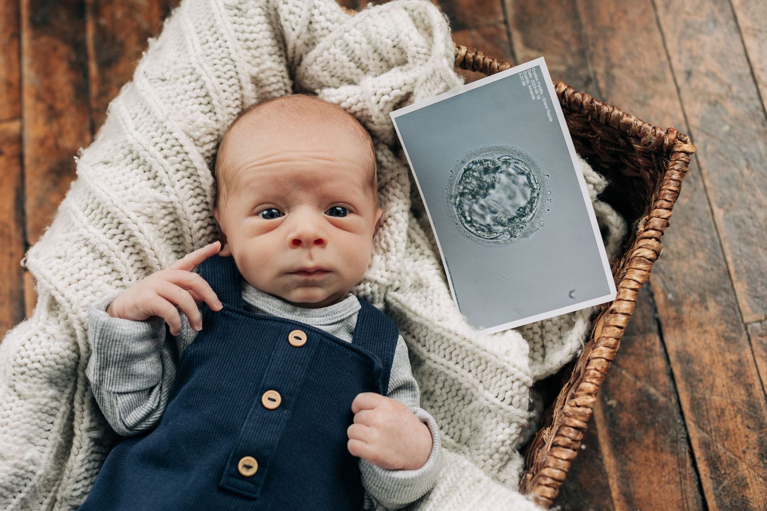 Baby in navy overalls lies on knit blanket next to gray passport holder on wooden floor.