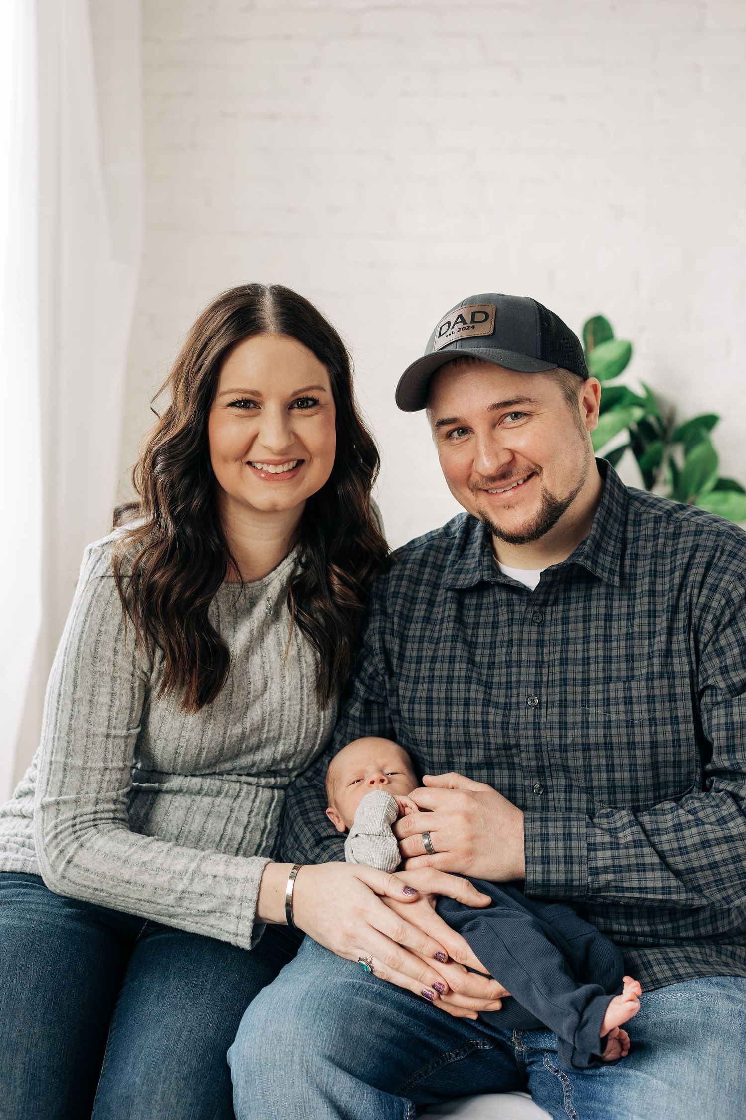 Family of three poses together on couch with newborn baby in casual indoor setting.
