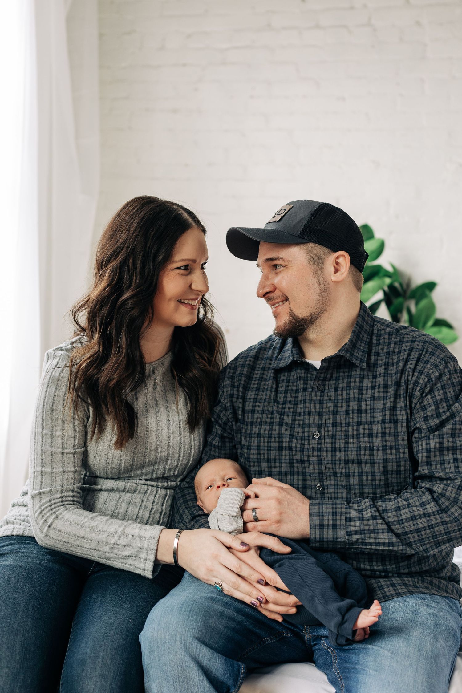 Couple smiles at each other while holding their newborn baby on gray couch.