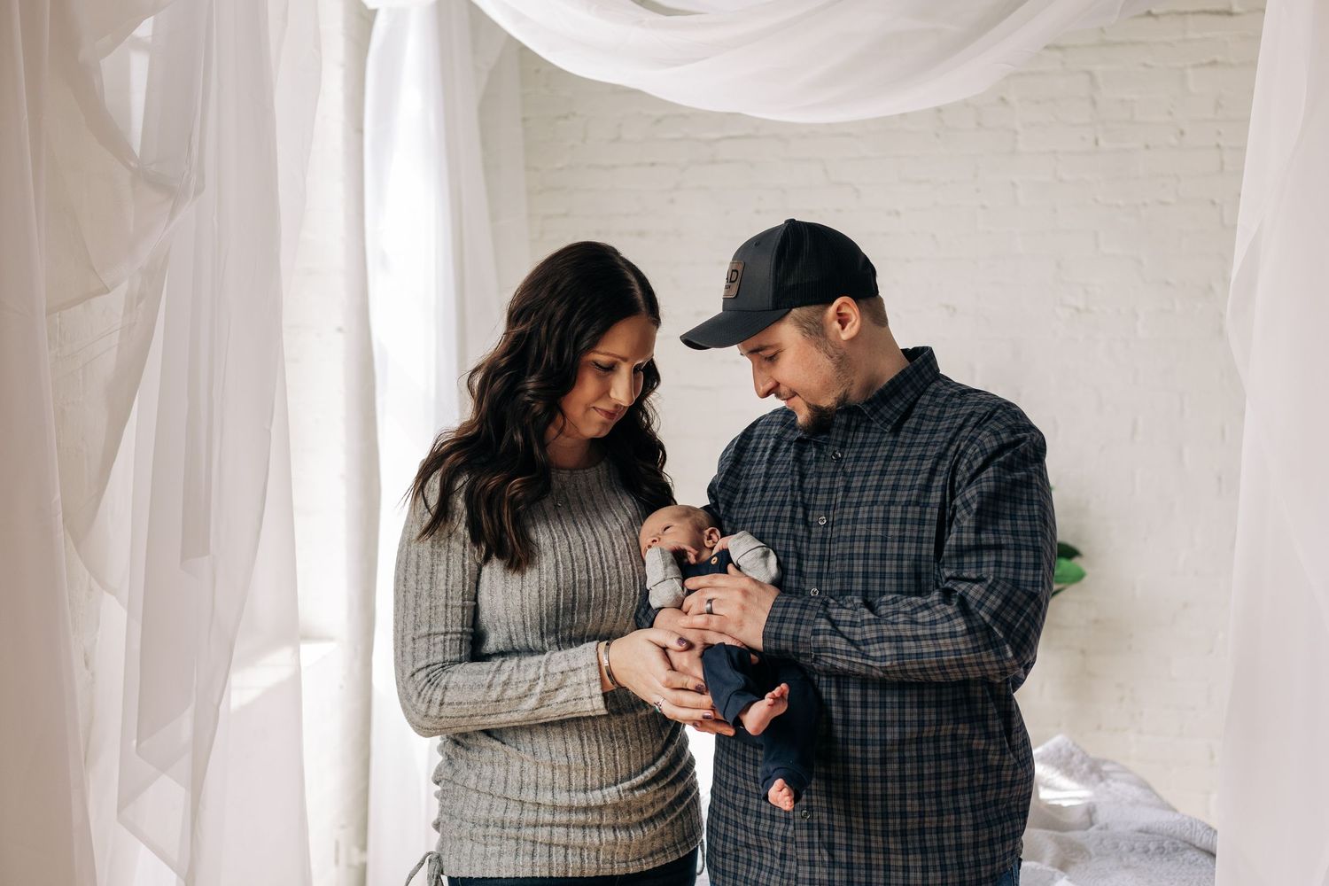 A couple in casual winter attire admiring something small together in a bright room with white curtains.