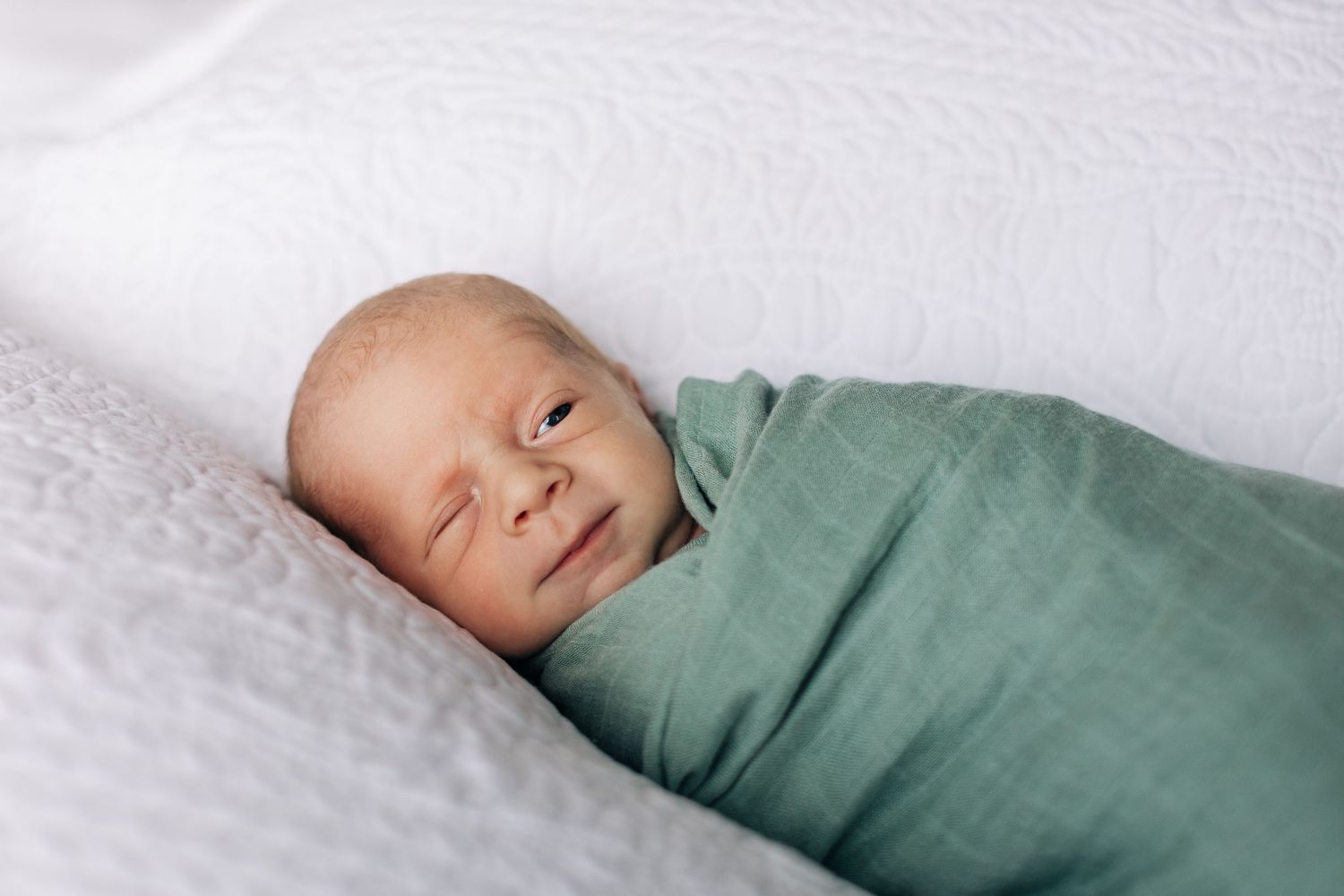 A newborn swaddled in mint green fabric resting on white bedding.