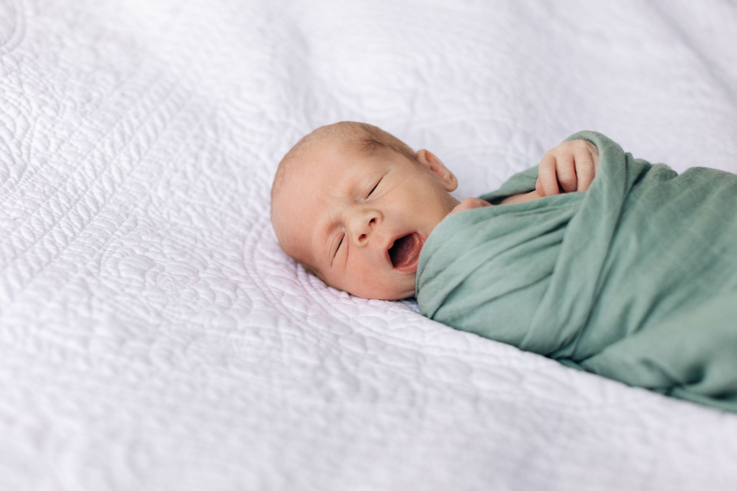A yawning infant wrapped in sage green swaddle on white textured bedding.