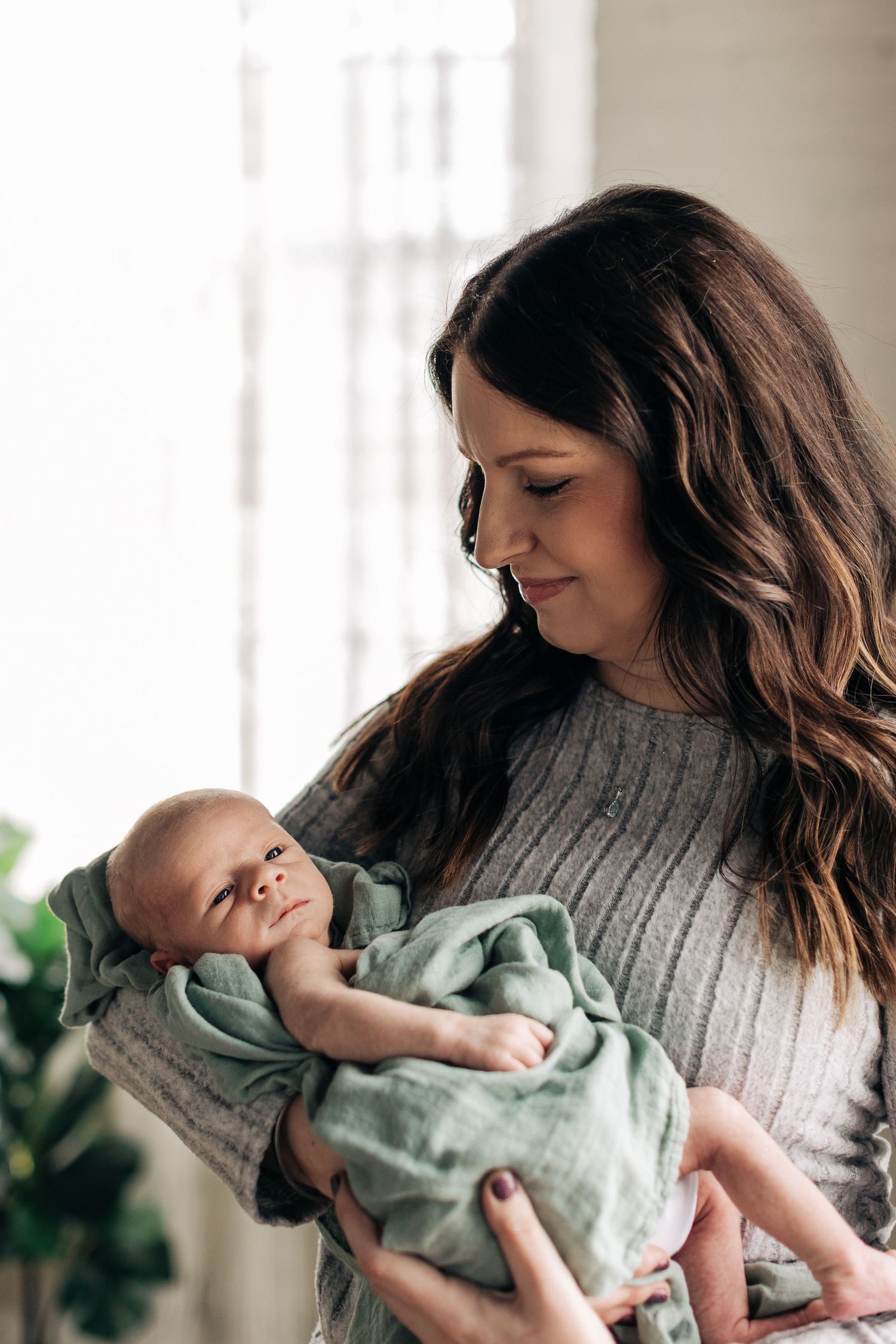 A woman in a gray sweater holds and gazes lovingly at a newborn baby wrapped in a mint green blanket.