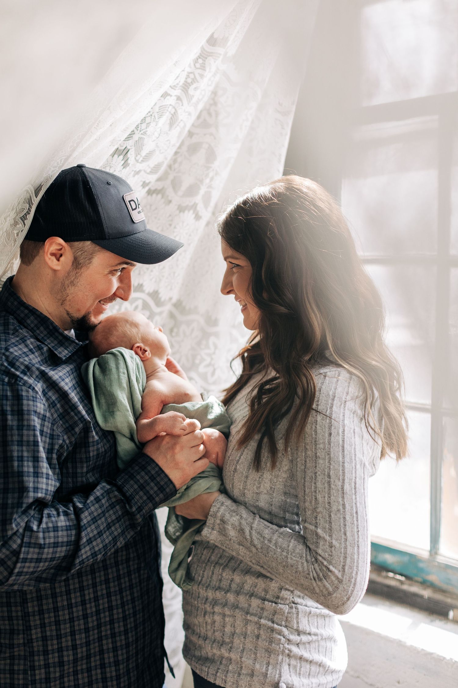 Parents gaze adoringly at their newborn baby while standing near a lace-curtained window.