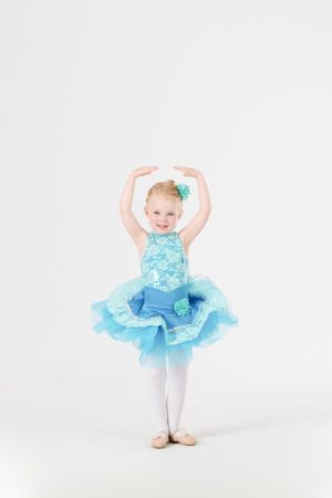 Young ballet dancer in light blue tutu costume with arms raised in graceful dance pose against white background.