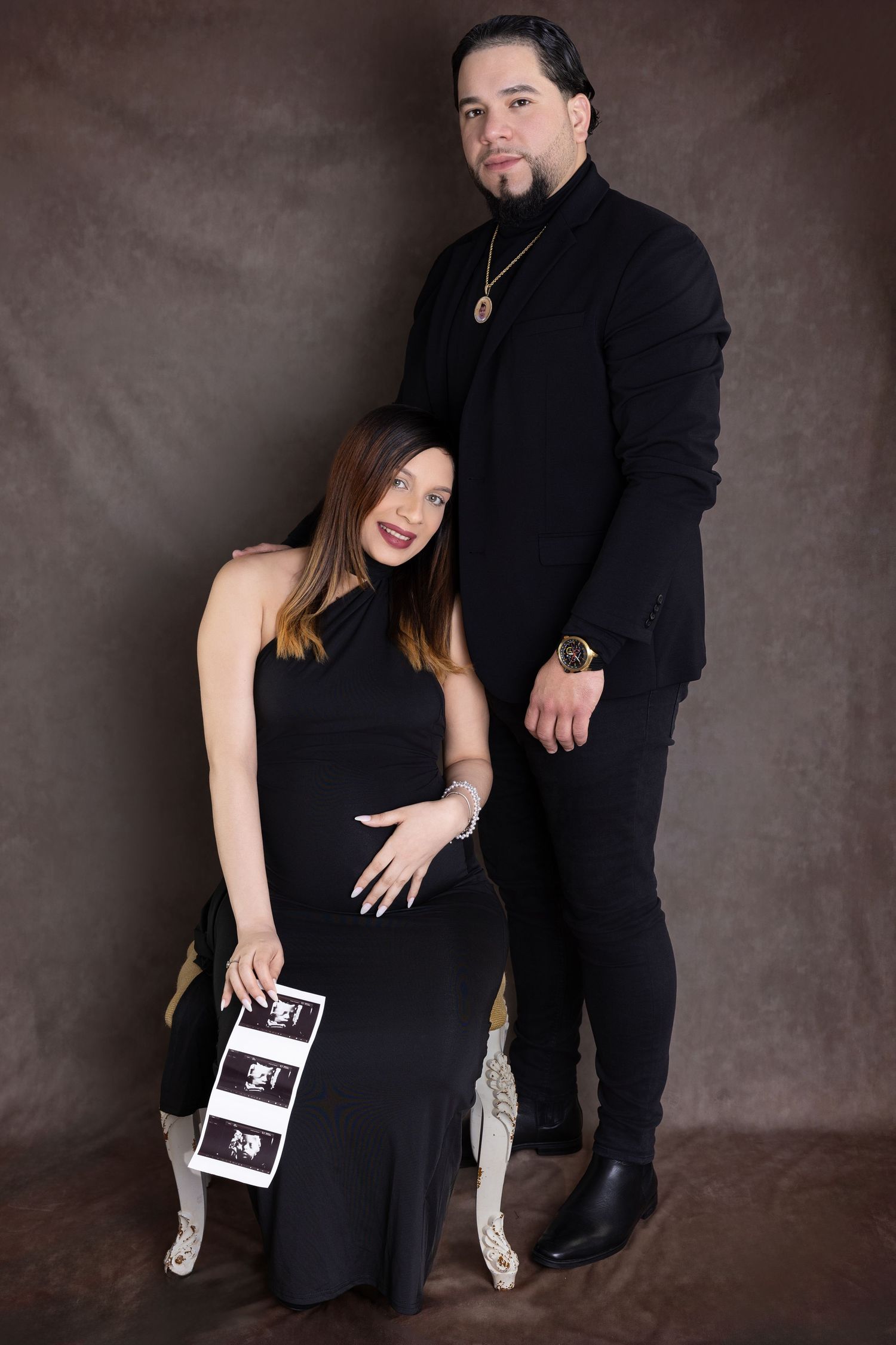 Couple in matching black outfits poses formally against dark backdrop for professional studio portrait.