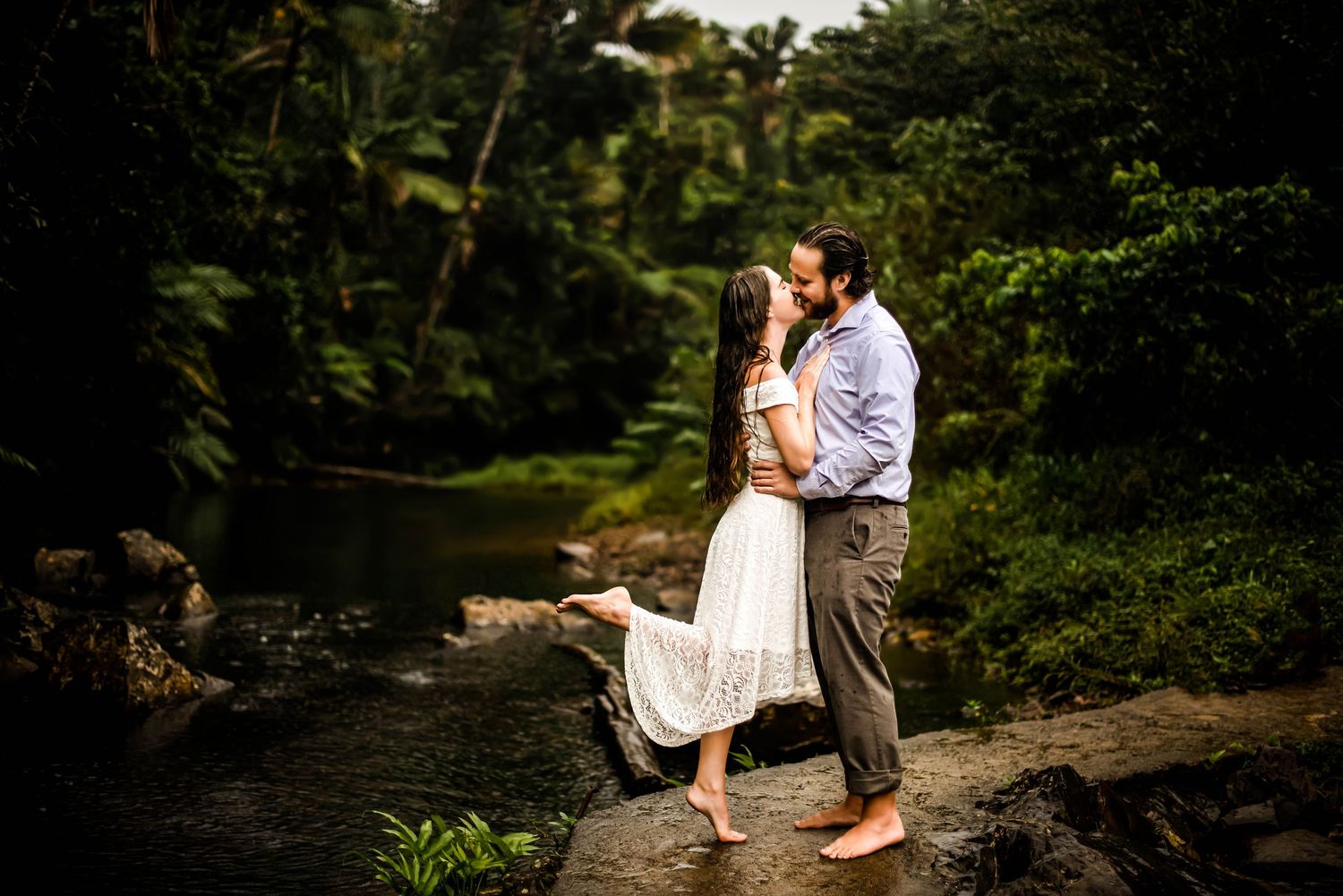 A romantic couple embraces on rocky terrain beside a lush forest stream.