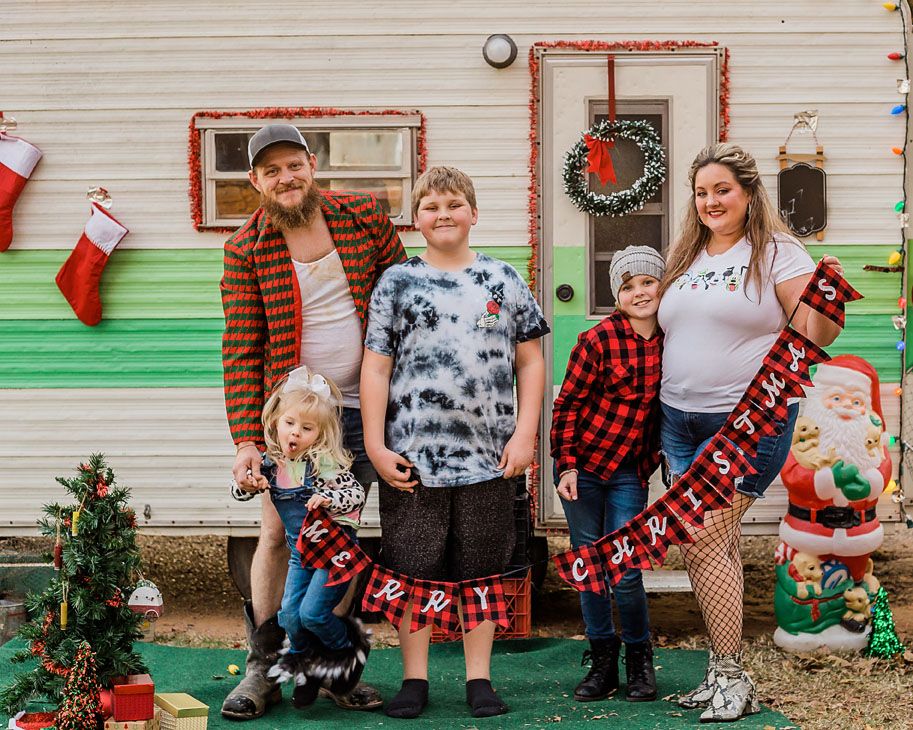 Family dressed in buffalo plaid poses for Christmas photo in front of decorated vintage camper.