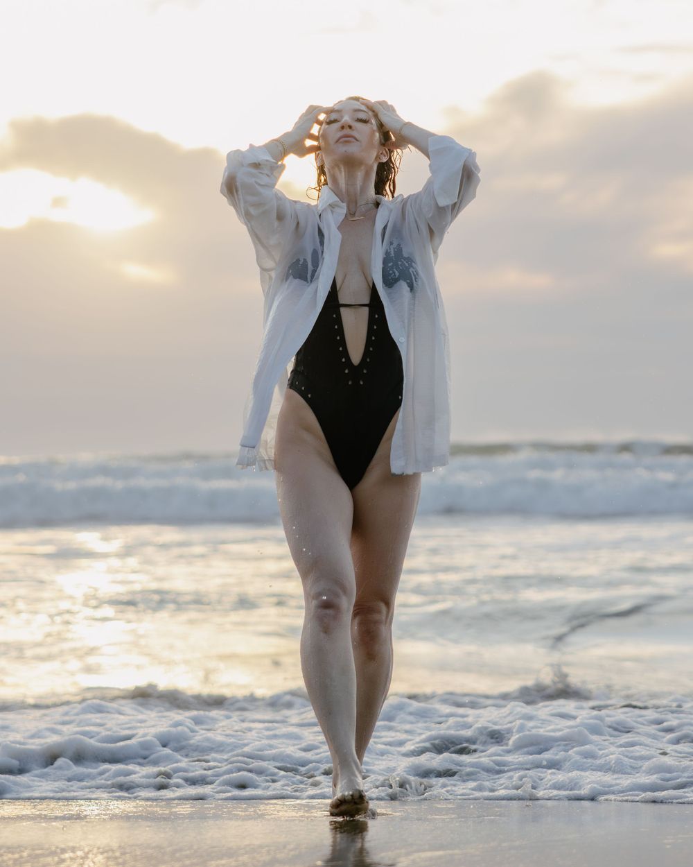 Figure in white oversized shirt and black swimsuit poses on beach at sunset with ocean waves in background.