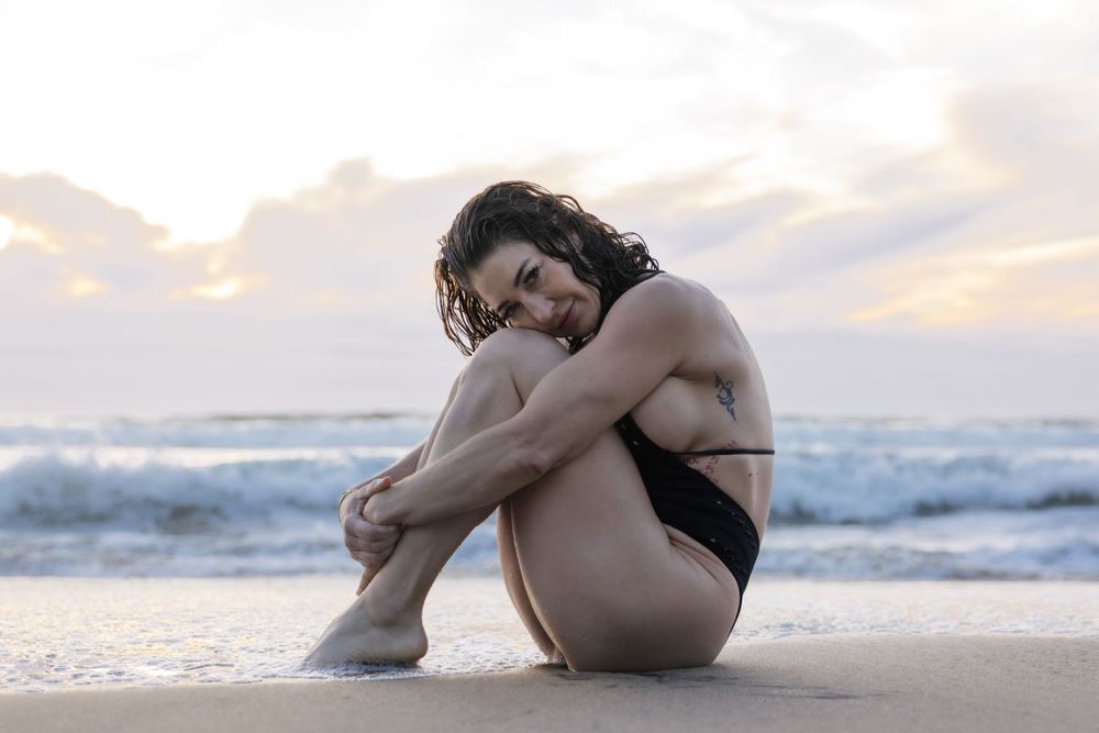 A person in a black swimsuit sits curled up on a sandy beach at sunset with waves crashing in the background.