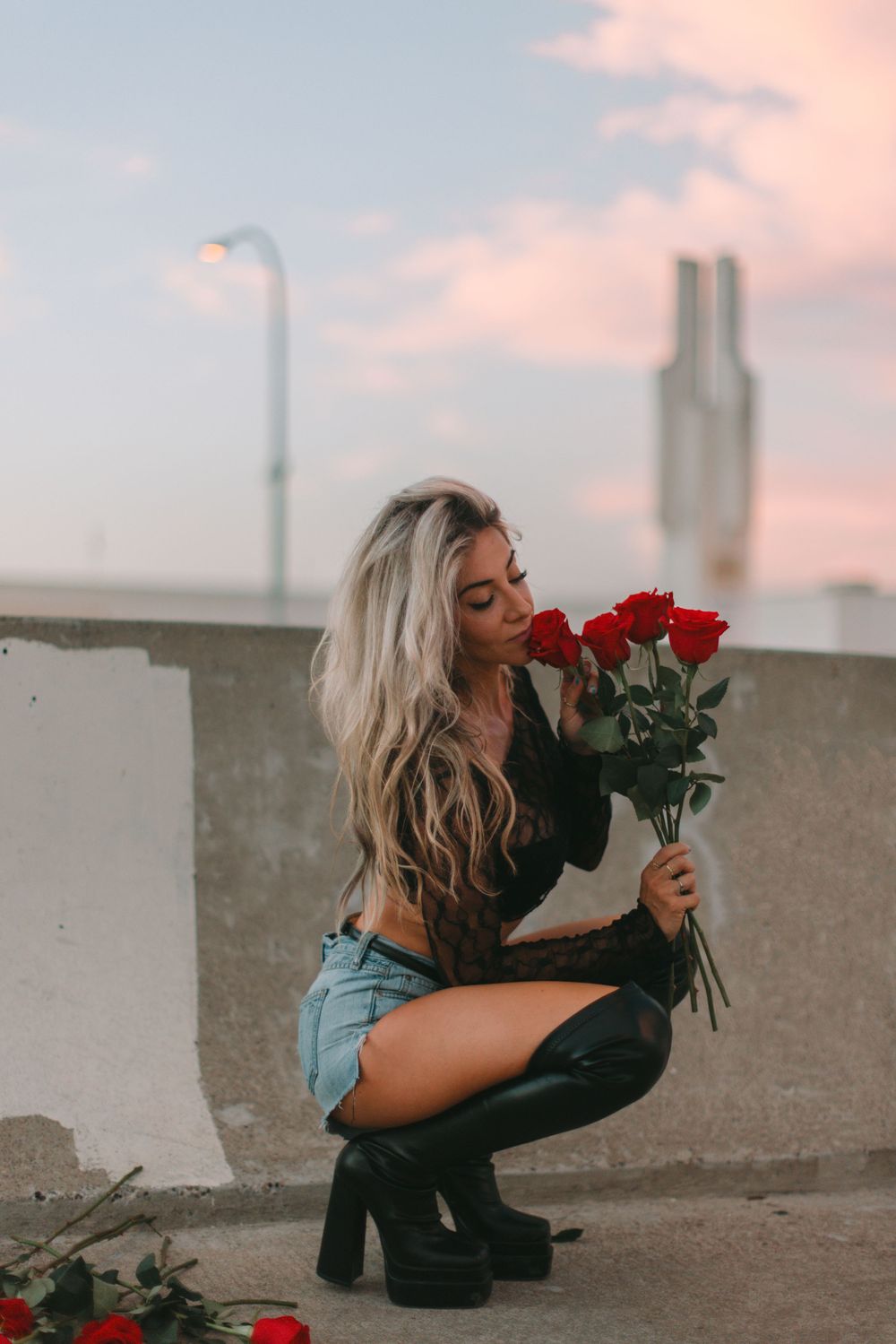 A person in black boots and denim shorts crouches down with a bouquet of red roses against a sunset sky backdrop.