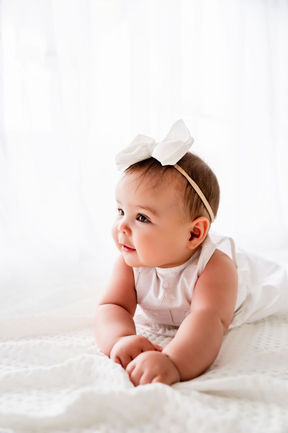 A sweet baby in white clothing lies on soft white bedding while smiling and playing in natural light.