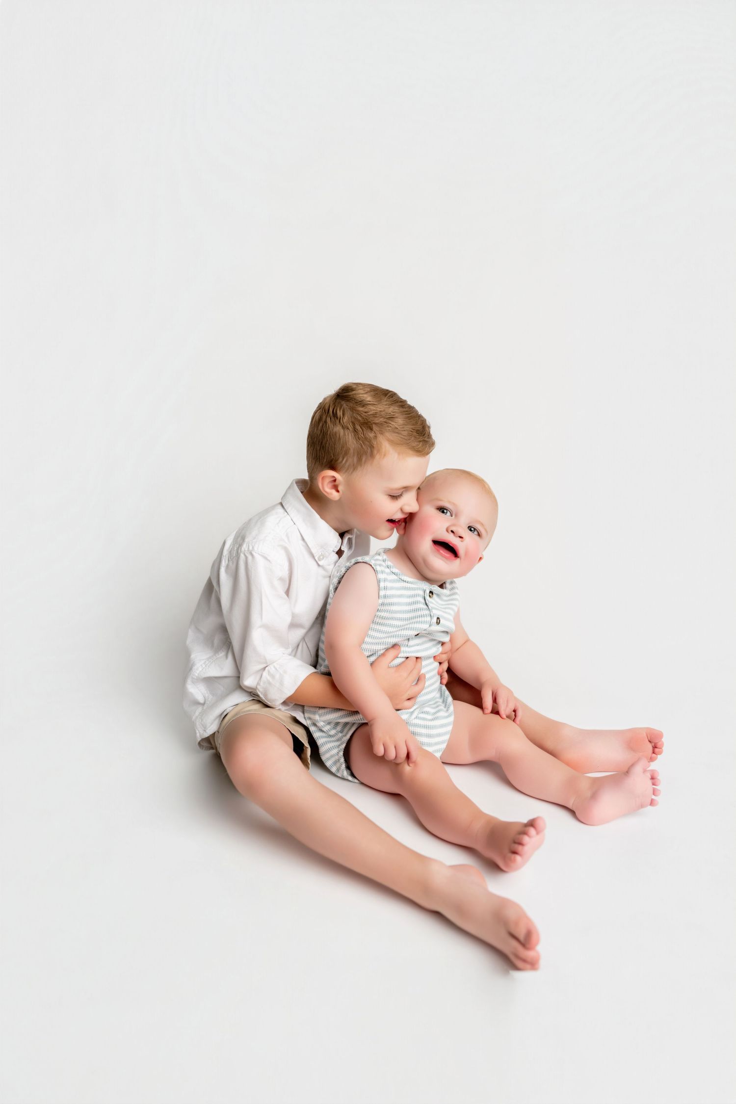 Older boy sitting on white background holding laughing baby sibling in striped onesie, sweet brotherly moment.