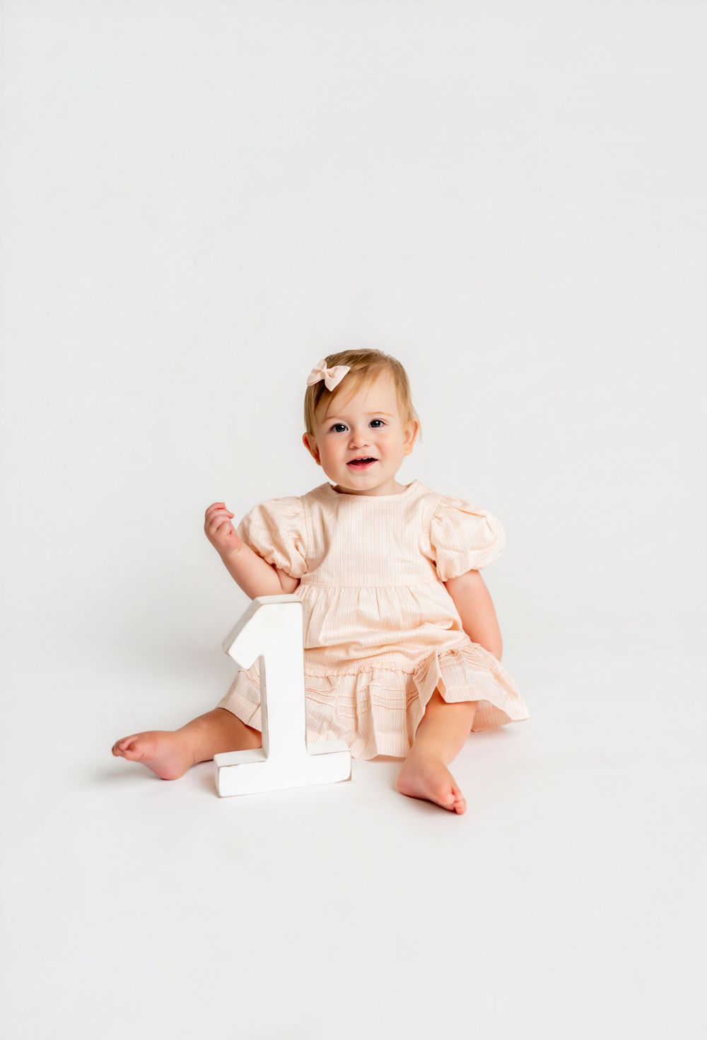 Smiling baby girl in peach dress sitting beside a white number one on white background.