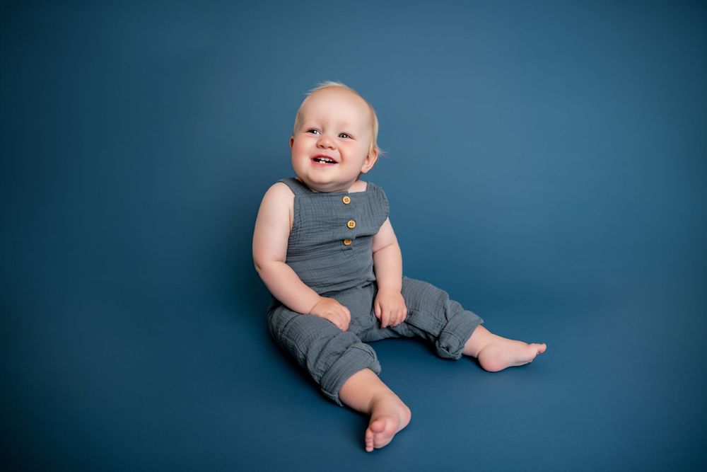 Smiling baby in gray romper sitting on blue background for a professional infant portrait photo.