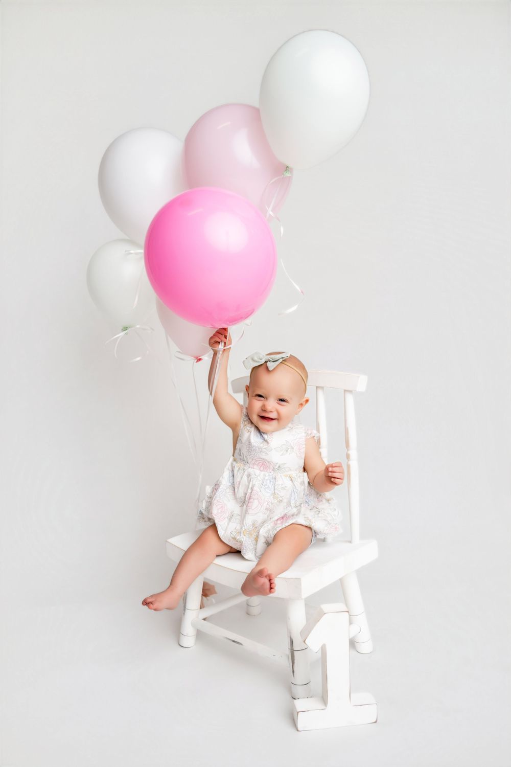 Happy baby girl in white dress sitting on chair holding pink and white balloons on white background.