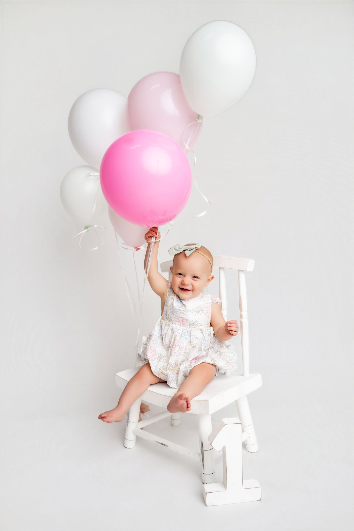 Happy baby girl in white dress sitting on chair holding pink and white balloons on white background.