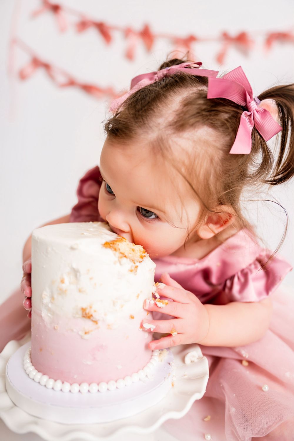 A small child in pink dress eagerly reaches for and tastes a delicate pink and white birthday cake.