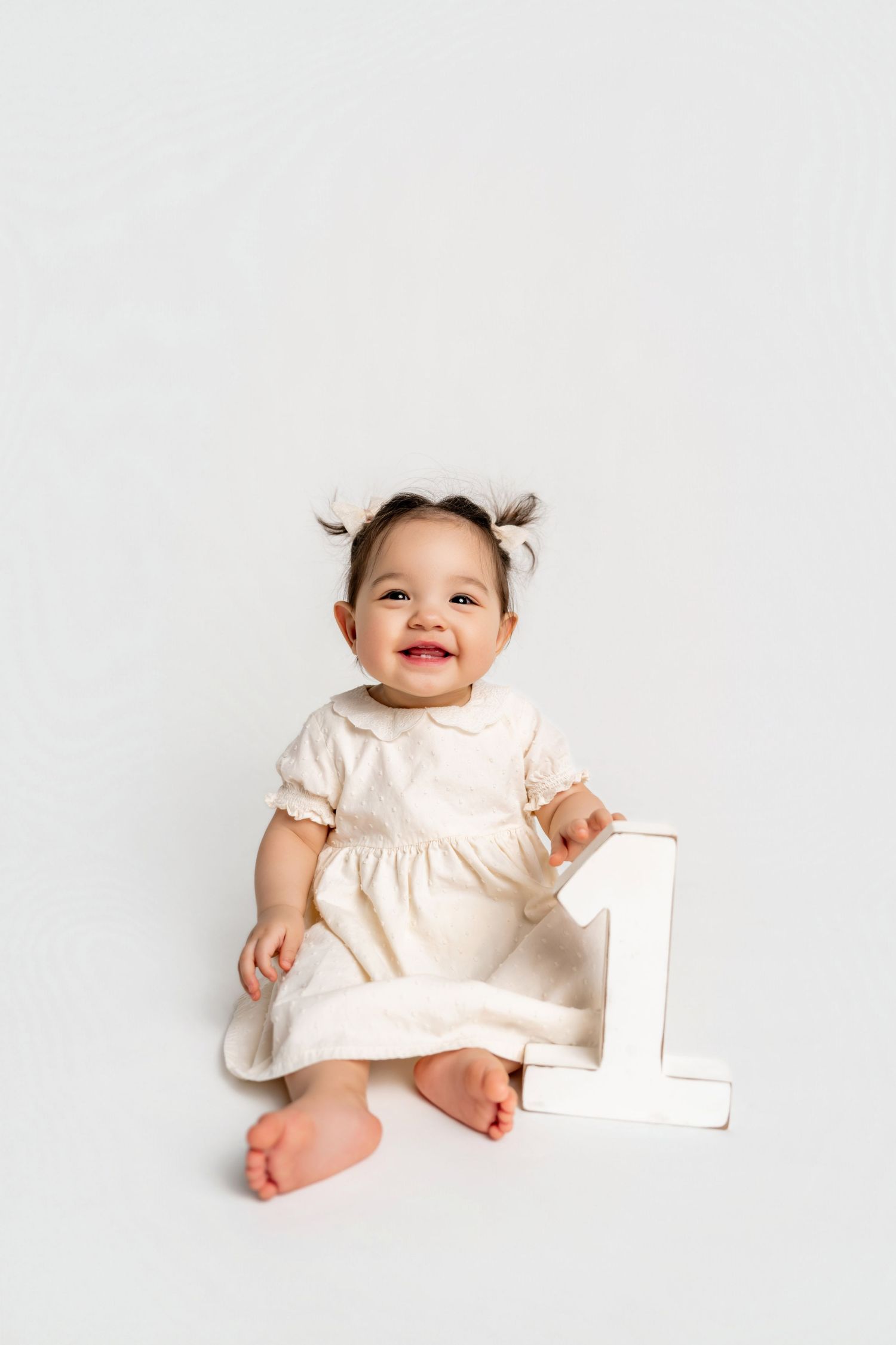 Smiling baby girl in cream dress sitting with number 1 sign on white background, celebrating first birthday.