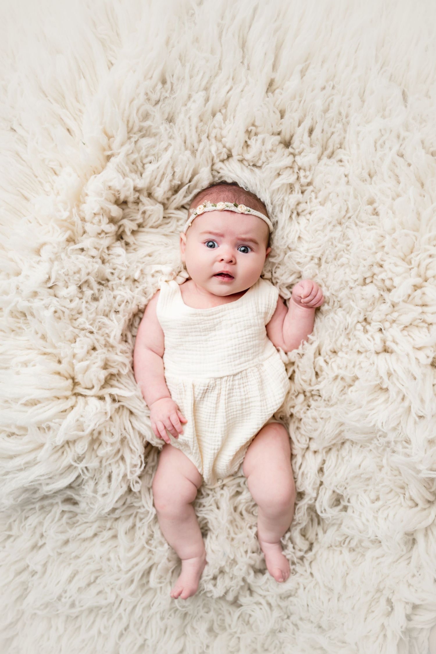 Adorable baby girl in cream romper and floral headband lying on fluffy white fur rug, looking up curiously.