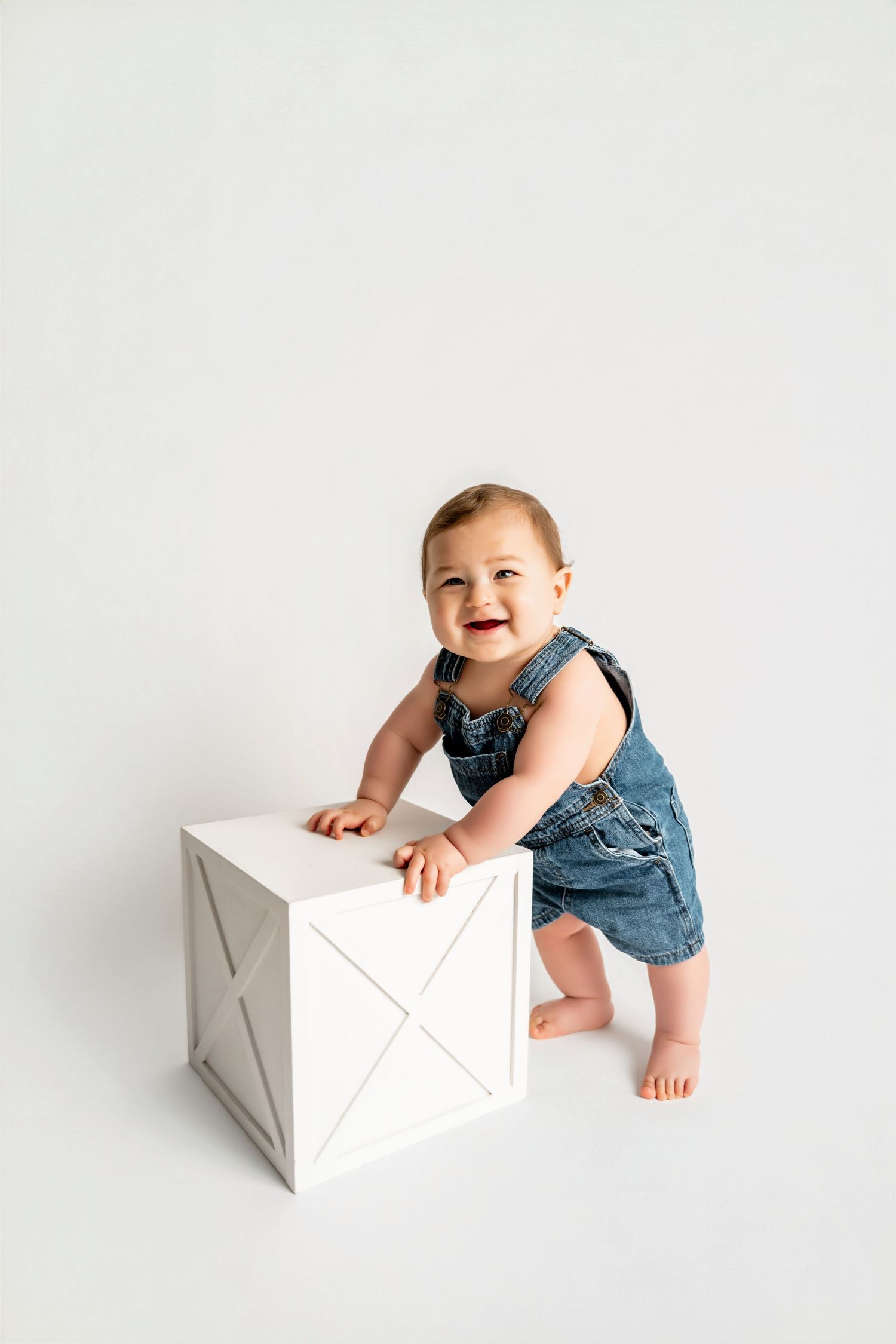 Smiling baby in denim overalls standing with support on a white wooden crate against a white background.