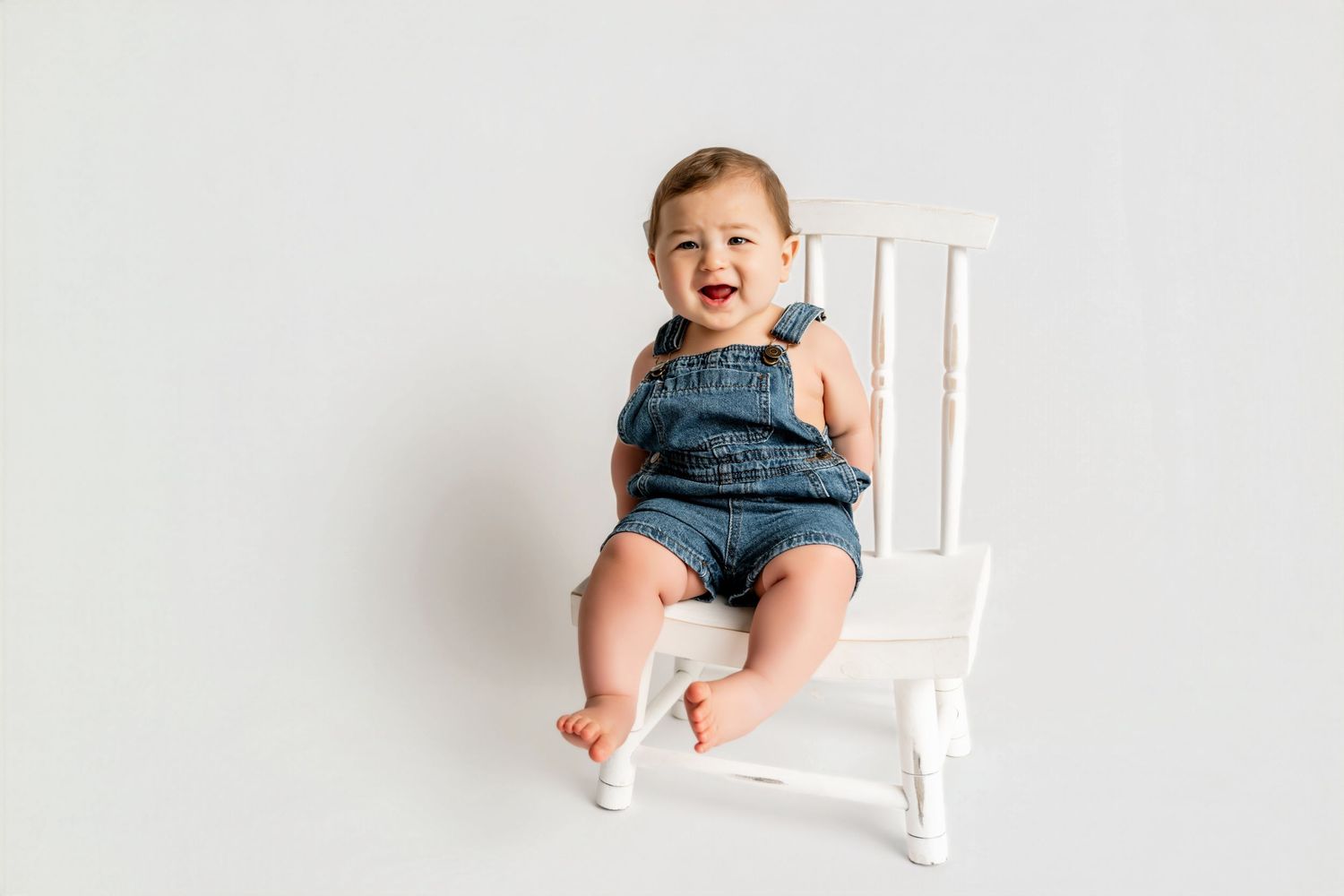 Smiling baby in denim overalls sitting on a small white chair against a white background.