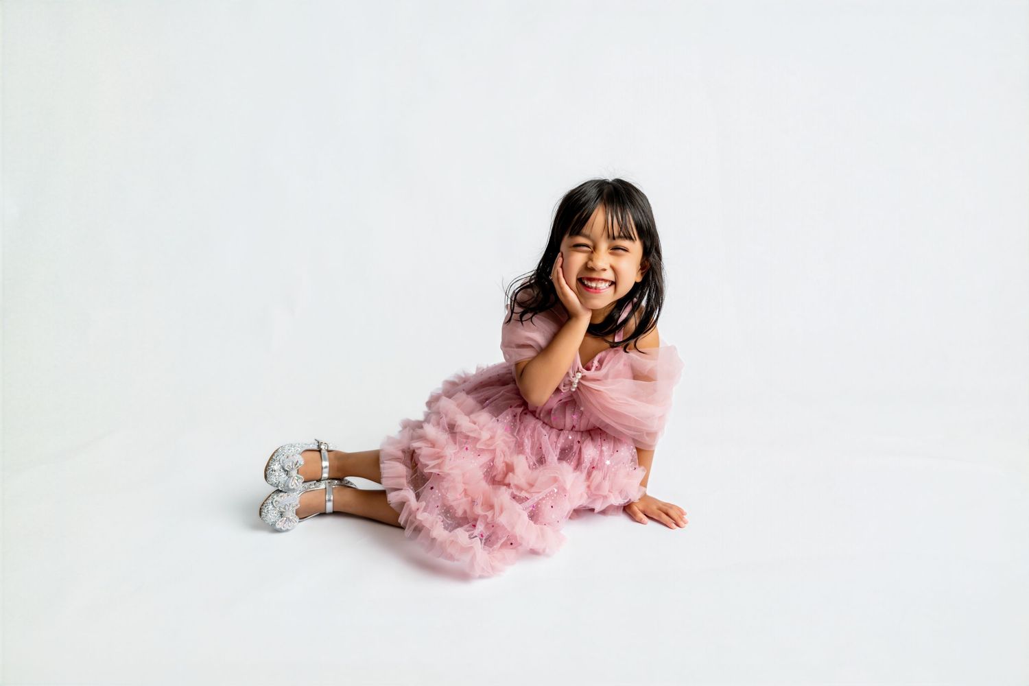 A young girl in a pink tulle dress and silver shoes sits smiling on a white background.