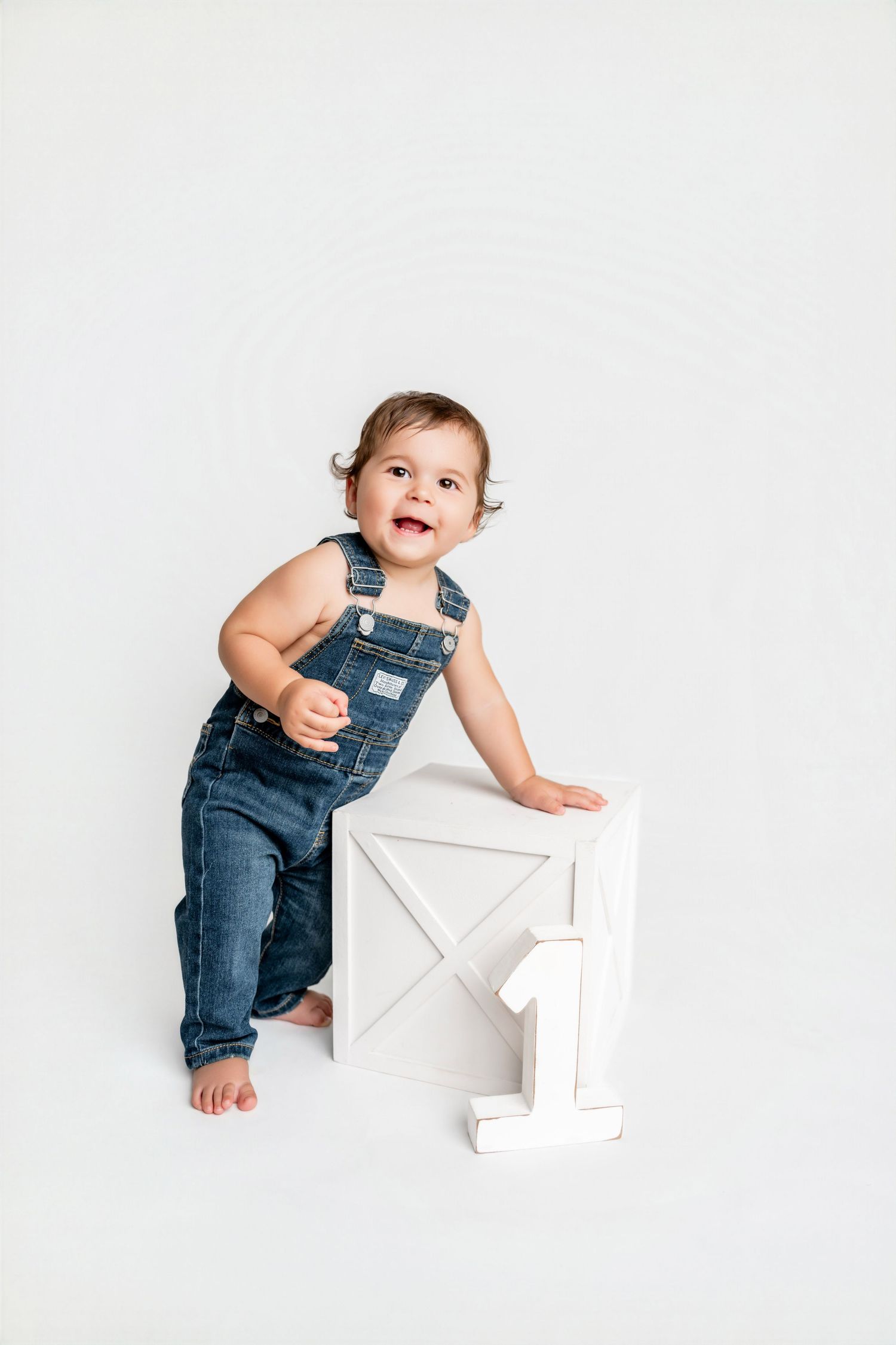 Smiling toddler in denim overalls standing next to a white wooden crate with the number 1, first birthday photo.