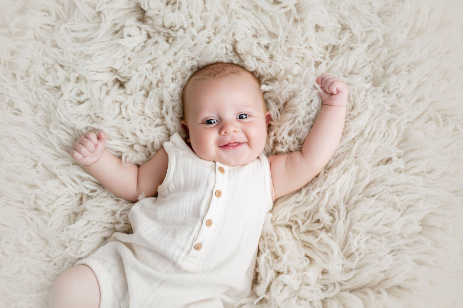Smiling baby in white romper lying on fluffy cream rug with arms raised, looking up at camera.
