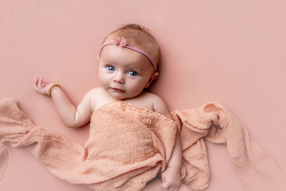 A newborn baby girl with blue eyes wrapped in a peach swaddle blanket, wearing a pink headband on a pink background.