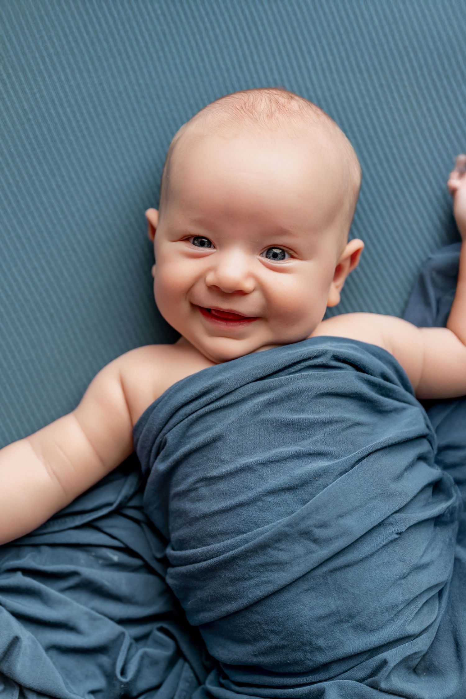 Smiling baby wrapped in a blue blanket lying on a blue background.